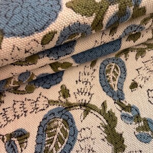 May include: Close-up of folded fabric with a floral pattern. The design features large blue flowers and green leaves on a cream-colored background. The fabric has a textured appearance, suggesting a woven material.