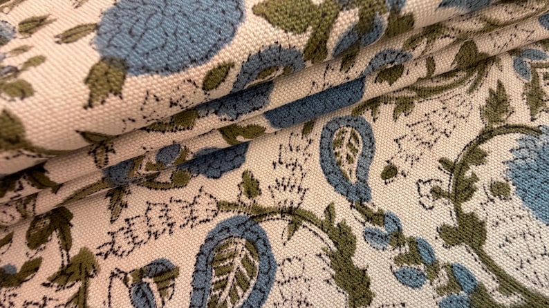 May include: Close-up of folded fabric with a floral pattern. The design features large blue flowers and green leaves on a cream-colored background. The fabric has a textured appearance, suggesting a woven material.