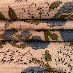 May include: Close-up of folded fabric with a floral pattern. The design features blue and green floral elements on a cream-colored background. The fabric appears to be a woven material, possibly cotton or linen.