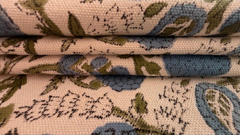 May include: Close-up of folded fabric with a floral pattern. The design features blue and green floral elements on a cream-colored background. The fabric appears to be a woven material, possibly cotton or linen.