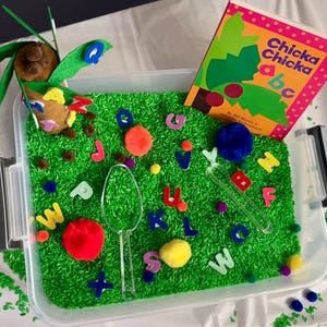 May include: A sensory bin filled with green dyed rice, colorful pom-poms, and alphabet letters. A clear plastic spoon and a children's book titled "Chicka Chicka abc" are also in the bin. A decorative palm tree is attached to the side.