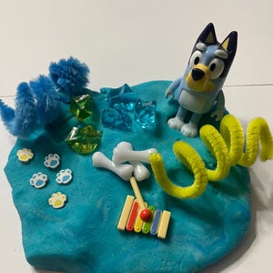 Pode incluir: Uma cena de plasticina azul com uma figura de Bluey, pompons azuis, pedras preciosas de plástico transparente, peças brancas em forma de osso, autocolantes de pegadas e um xilofone de brinquedo com uma maça vermelha. Um limpador de cachimbo amarelo está enrolado perto.