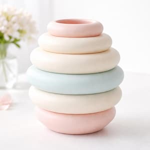 May include: A pastel-colored stacking toy with six rounded rings in shades of pink, cream, and blue. The rings decrease in size, creating a pyramid shape. The toy is likely made of ceramic or a similar material.