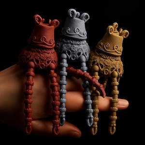 May include: Three decorative, abstract sculptures in shades of red, gray, and gold. Each sculpture has a bulbous head with ornate detailing and long, segmented legs. The sculptures are held in a hand against a black background.