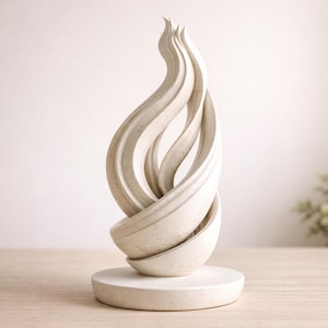 May include: A cream-colored abstract sculpture with a flame-like design. The sculpture features intertwined, flowing shapes that rise from a circular base. The piece has a textured surface and is displayed on a light-colored wooden surface.
