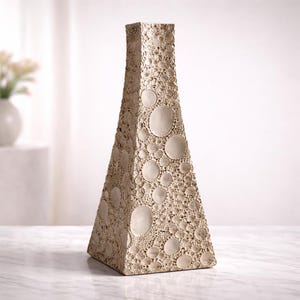 May include: A tall, beige, textured vase with a geometric, triangular shape. The surface is covered in a pattern of varying-sized circles, creating a unique, tactile appearance. The vase is a decorative home accent.