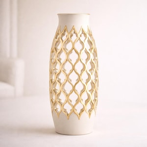 Puede incluir: Un jarrón alto de color blanco roto con un patrón geométrico dorado. El jarrón tiene un cuello estrecho y una base ancha. El patrón dorado es un diseño abierto y repetitivo que cubre la mayor parte de la superficie del jarrón. El borde del jarrón también está adornado con oro.