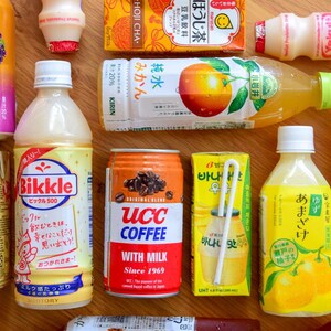 May include: An assortment of Japanese beverages, including canned coffee, juice boxes, and bottled drinks. The products feature colorful labels with Japanese text and graphics, showcasing various flavors and brands. The arrangement is on a wooden surface.