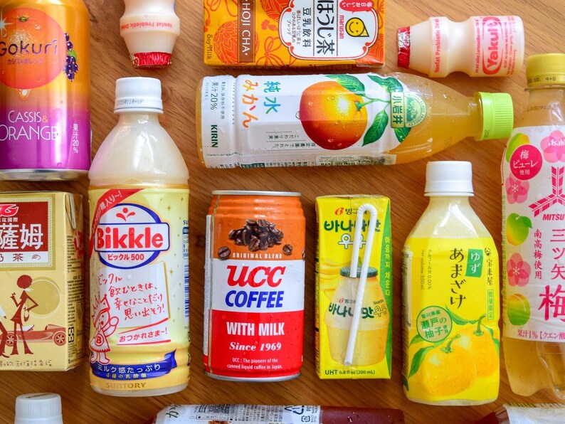 May include: An assortment of Japanese beverages, including canned coffee, juice boxes, and bottled drinks. The products feature colorful labels with Japanese text and graphics, showcasing various flavors and brands. The arrangement is on a wooden surface.