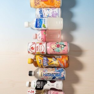 May include: A collection of various Japanese beverages, including soda, milk tea, and fruit-flavored drinks. The bottles and cans are arranged vertically, showcasing colorful labels with Japanese text and illustrations. The selection includes strawberry milk, soda, and other unique flavors.