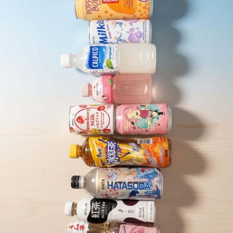 May include: A collection of various Japanese beverages, including soda, milk tea, and fruit-flavored drinks. The bottles and cans are arranged vertically, showcasing colorful labels with Japanese text and illustrations. The selection includes strawberry milk, soda, and other unique flavors.