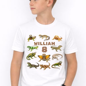 Könnte beinhalten: Weißes T-Shirt mit bunten Eidechsen-Illustrationen. Der Name "WILLIAM" und die Zahl "8" sind in brauner, strukturierter Schrift gedruckt. Ein lässiges Kleidungsstück für den Alltag.