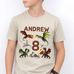 Puede incluir: Camiseta beige con el nombre "ANDREW" en letras marrones y el número "8". La camiseta está decorada con ilustraciones de dragones coloridos, algunos escupiendo fuego.
