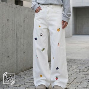 May include: White denim pants with embroidered fruit designs, including an orange, apple, avocado, and strawberry. The pants have a wide leg and a classic five-pocket design. The model is wearing a gray sweatshirt.