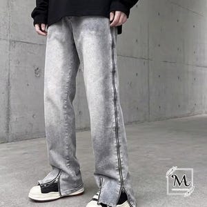 May include: Light grey denim trousers featuring black zippers along the sides and ankles. The trousers have a relaxed fit and are paired with black and white trainers. The backdrop is a concrete wall.