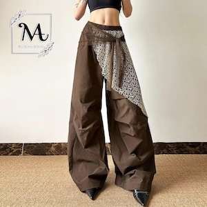 May include: Brown wide-leg trousers with a distinctive lace overlay. The trousers have a high waist and a flowing, oversized fit. The lace detail adds a touch of elegance to the casual design. The trousers are paired with black pointed-toe shoes.