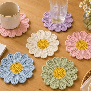 May include: A set of six round, flower-shaped crochet coasters in pastel pink, light purple, cream, light blue, and sage green. Each coaster features a bright yellow circular center with twelve rounded petals radiating outward in a daisy pattern.