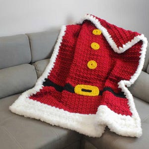 May include: A red crocheted blanket designed to resemble Santa Claus's suit. The blanket features yellow buttons, a black belt with a yellow buckle, and white fluffy trim. The blanket is draped over a gray sofa.