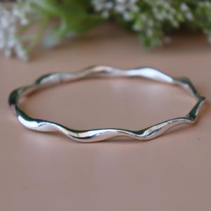 May include: A silver bangle bracelet with a flowing, wavy design. The bracelet has a smooth, polished finish and an irregular shape. The jewellery is displayed on a light pink surface.