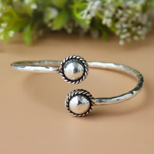 May include: A silver-toned cuff bracelet with a hammered texture. The bracelet features two large, round, polished silver beads, each encircled by a twisted rope design. The bracelet is open, with the beads at each end.