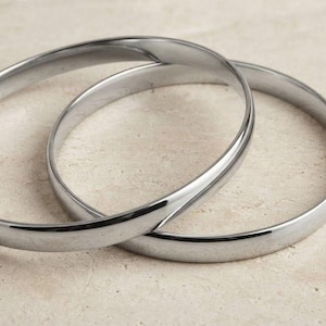 May include: Two polished silver-tone metal bangle bracelets with a smooth, rounded surface. The two circular bangles are interlocked, creating a continuous loop design with a reflective, high-shine finish on the exterior metal surface.