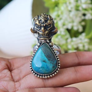 Peut inclure: Bague en argent ornée d'une tête de démon en laiton et d'une pierre turquoise en forme de goutte. La pierre est encadrée d'une bordure argentée perlée. La bague est posée sur un fond flou.