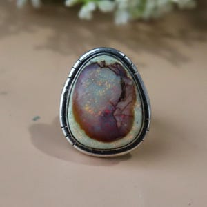 May include: A silver ring featuring a large, oval-shaped gemstone. The stone has a mix of colors, including brown, red, and green, with a cracked pattern. The ring's setting is a simple, polished silver band.