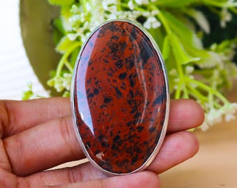 May include: Oval-shaped, polished red and black gemstone set in a silver-colored metal ring. The stone has a unique pattern of dark spots and swirls. The ring is held in a hand, with a blurred background of green leaves and white flowers.