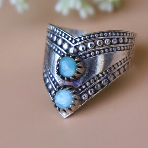 May include: A wide, silver-toned ring featuring two light blue, oval-shaped gemstones. The ring has a textured, geometric design with small, circular accents and a V-shaped front. The gemstones are set in a decorative, serrated bezel.