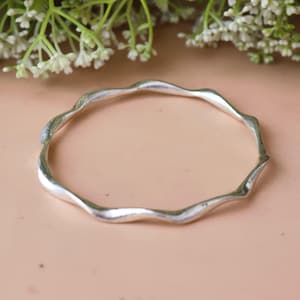 May include: A silver-colored bangle bracelet with a wavy, organic design. The bracelet has a smooth, polished finish and a circular shape. The background is a soft, muted pink, with a sprig of white flowers at the top.