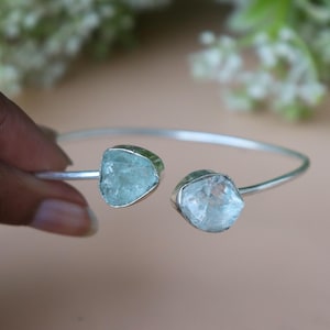 May include: A silver-toned bracelet featuring two light blue aquamarine stones. The stones are irregularly shaped and set in a silver bezel. The bracelet is open-ended, allowing for easy wear.