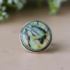May include: A round silver ring featuring a large, iridescent opal stone. The opal displays a mix of green, blue, and brown colors with a cracked pattern. The ring has a simple, polished silver band.