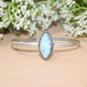 May include: A silver bracelet featuring a light blue, oval-shaped gemstone. The gemstone is set in a twisted rope-like border. The bracelet has a simple, polished band. The background includes white flowers and a neutral-toned surface.