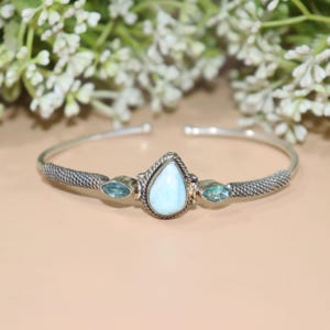 May include: A silver cuff bracelet featuring a pear-shaped, light blue gemstone in the center, flanked by two smaller, oval-shaped blue gemstones. The bracelet has a textured design on the sides and is set against a neutral background.