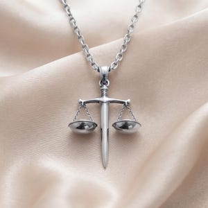 May include: A silver-toned metal necklace featuring a pendant shaped like a sword integrated with a set of balanced scales. The pendant hangs from a matching silver-toned link chain, showcasing a polished finish and detailed metalwork throughout.