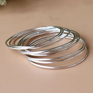 May include: A stack of silver bangles. These thin, round bracelets are made of shiny metal and are stacked together. The bangles are displayed on a neutral-colored surface.