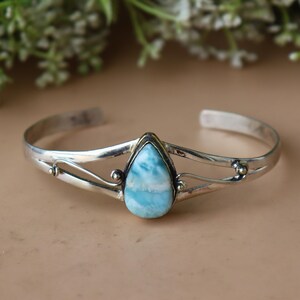 May include: A silver cuff bracelet with a teardrop-shaped, light blue gemstone. The gemstone is set in a gold-colored bezel. The bracelet has decorative silver accents on either side of the gemstone.