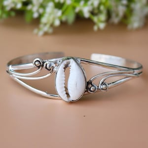 May include: A silver cuff bracelet featuring a white seashell centerpiece. The bracelet has an open design with decorative swirls and curves. The seashell is oval-shaped with a natural, textured surface. The bracelet is displayed on a neutral background.