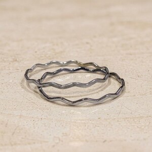 May include: Two thin, circular metal rings with a continuous wavy or zig-zag pattern along the entire circumference. The rings have a dark, metallic finish with a subtle textured surface, appearing as stacked bands with a uniform, repeating design.