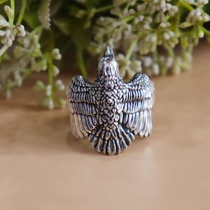 May include: A silver-colored ring shaped like a bird with outstretched wings. The ring has detailed feather textures and a pointed beak. The bird's wings are spread wide, creating a bold and eye-catching design. The ring is a piece of jewelry.