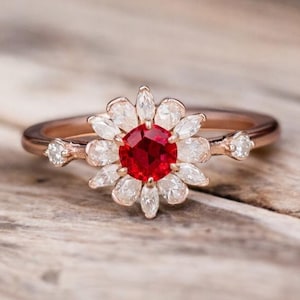 May include: A rose gold ring with a central, round, bright red gemstone encircled by white, petal-shaped stones. The ring's band is slender, and small accent stones are set on either side of the central design. The ring is displayed on a wooden surface.