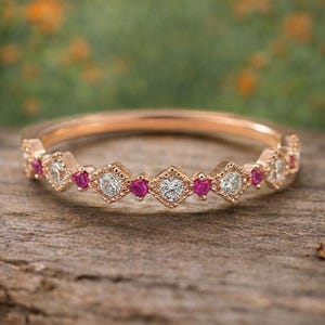 Può includere: Un anello in oro rosa con un motivo ripetuto di diamanti e piccole pietre preziose rosse. L'anello è appoggiato su una superficie di legno con uno sfondo sfocato verde e arancione.