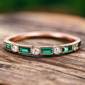 May include: Rose gold ring with alternating emerald-cut green and round white gemstones. The ring is displayed on a wooden surface with a blurred green backdrop. The design of the ring is elegant and striking.