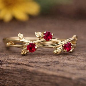May include: A gold-colored ring with a nature-inspired design. The ring features three red gemstones set among delicate leaves and branches. The ring is displayed on a wooden surface, with a blurred yellow flower in the background.
