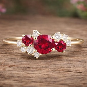 May include: A gold ring with a cluster of red and white gemstones. The ring's design features a central red stone surrounded by smaller red and white stones. The band is slender, and the background is a wooden surface with pink flowers.