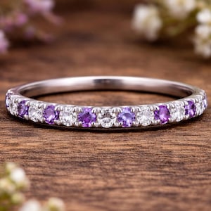 May include: A silver ring featuring alternating purple and white gemstones. The ring is set against a blurred floral background and a wooden surface, highlighting its intricate design and sparkle.