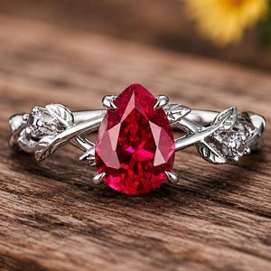 Op de afbeelding: Een zilverkleurige ring met een centrale peervormige rode edelsteen in een zetting. De band is ontworpen met een metalen wijnrank- en bladmotief, geaccentueerd door kleine, heldere, rondgeslepen stenen in het metalen bladwerk.