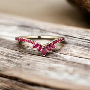 Op de afbeelding: Een goudkleurige ring met een gebogen ontwerp, bezet met kleine rode edelstenen langs de band en grotere, marquise-geslepen rode stenen die een centraal bloem- of bladachtig patroon vormen. De ring wordt gepresenteerd op een houten oppervlak.