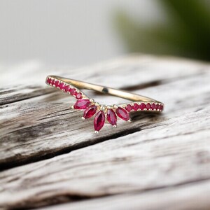 Op de afbeelding: Een goudkleurige ring met een gebogen ontwerp, bezet met kleine, helderrode edelstenen. De ring heeft een centrale cluster van marquise-geslepen robijnen, wat een vleugje elegantie toevoegt. De ring wordt gepresenteerd op een verweerd houten oppervlak.