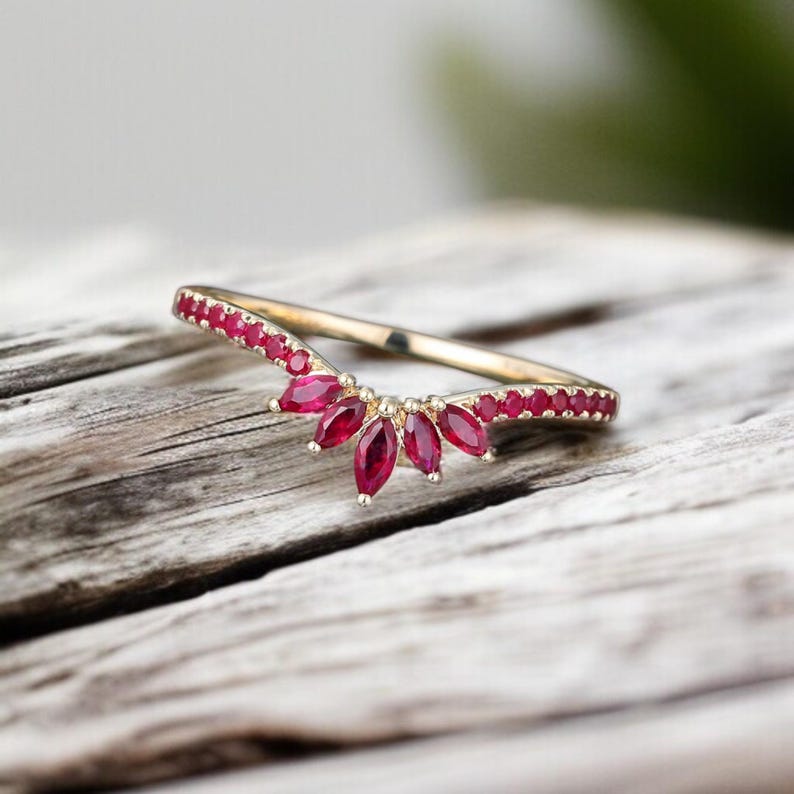 Op de afbeelding: Een goudkleurige ring met een gebogen ontwerp, bezet met kleine, helderrode edelstenen. De ring heeft een centrale cluster van marquise-geslepen robijnen, wat een vleugje elegantie toevoegt. De ring wordt gepresenteerd op een verweerd houten oppervlak.